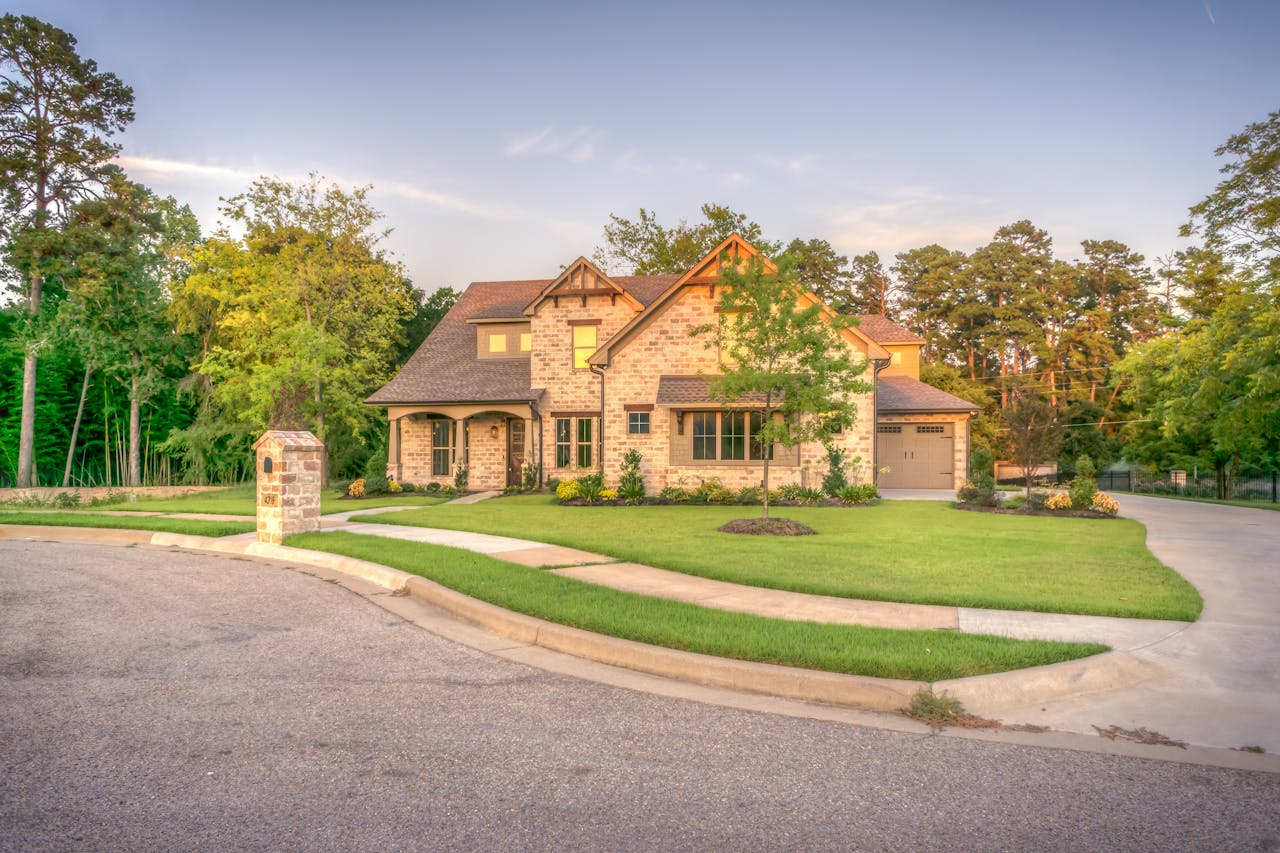 gallery-img-06 Elegant stone family home with manicured lawn, trees, and driveway on a sunny day.