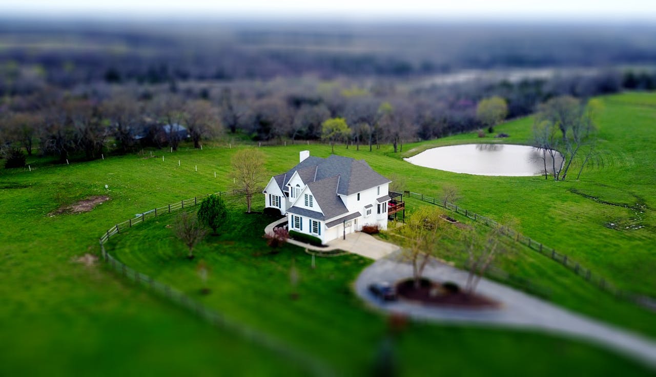 programs-01 Aerial shot of a charming rural home surrounded by lush green fields and a tranquil pond.